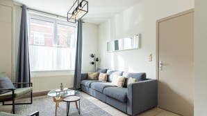 Ein modernes Wohnzimmer mit einem grauen Sofa, einem Glastisch und einem großen Fenster mit Vorhängen.