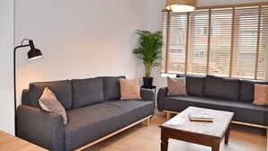 Un salon moderne avec deux canapés gris, une table basse en bois, un lampadaire et une grande fenêtre avec des stores.