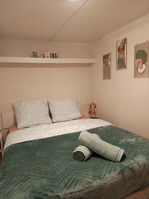 Un dormitorio con cama, almohadas, una toalla doblada, una mesita de noche, un estante con adornos y una pared con obras de arte enmarcadas.