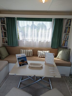 A cozy room with a sofa, a table with a placemat, a framed certificate, and bookshelves with books. - Escape Baie de Somme: Mobil home _ Vironchaux (Vironchaux)