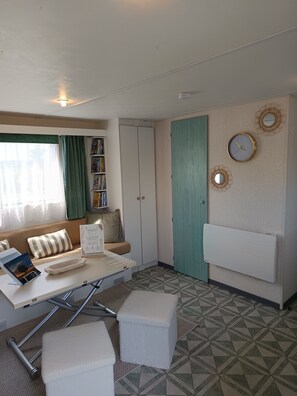 A room with a sofa, a small table, a radiator, a bookshelf, and a door. - Escape Baie de Somme: Mobil home _ Vironchaux (Vironchaux)