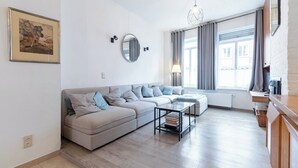 A modern living room with a sectional sofa, a round mirror, a glass coffee table, and a view of the city through large windows. - Spacious Bruges House with Free Parking (Brugge)