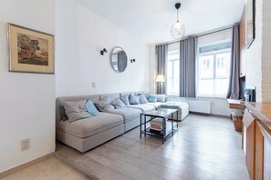 Ein modernes Wohnzimmer mit einem Ecksofa, einem runden Spiegel, einem Glastisch und Blick auf die Stadt durch große Fenster.