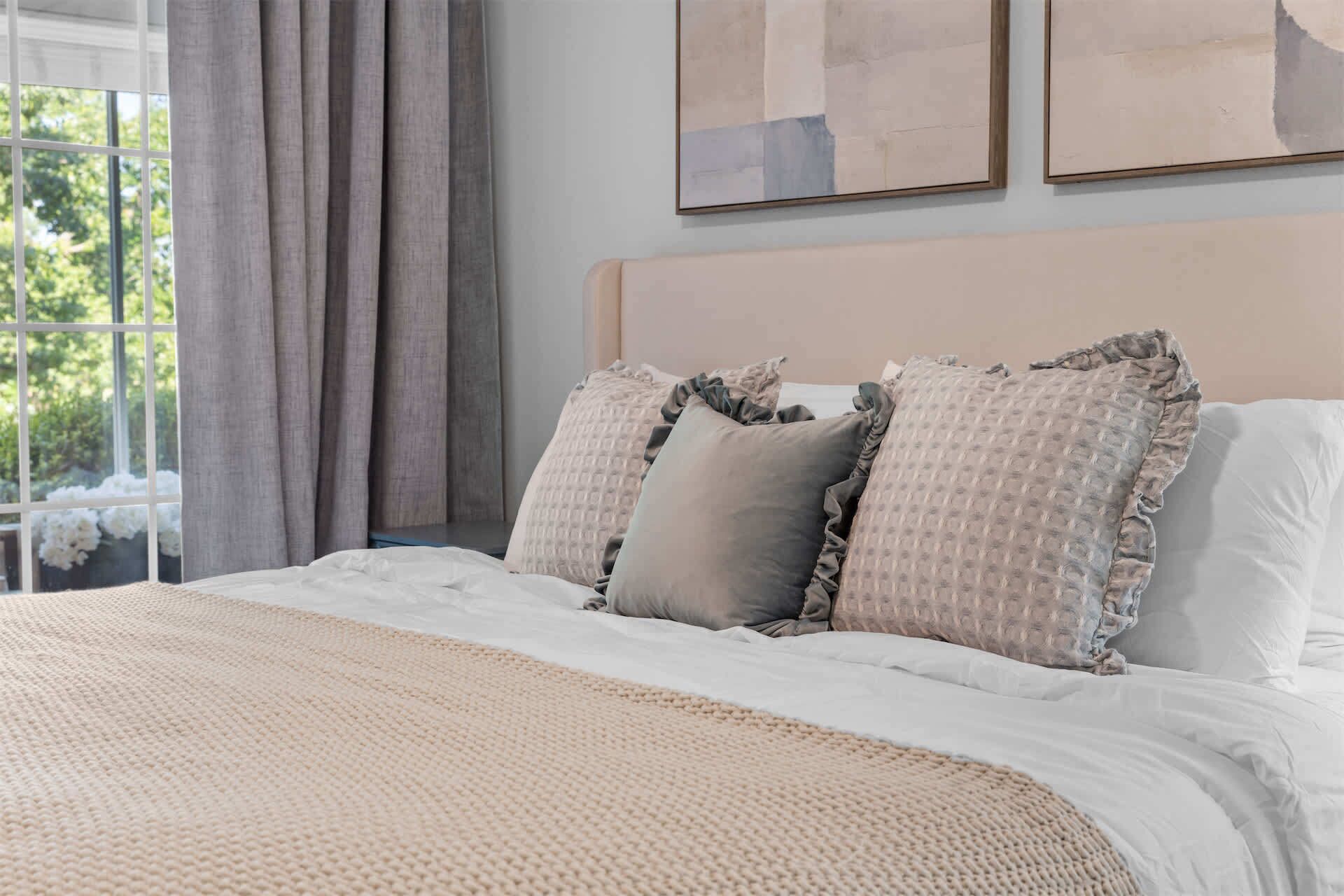 A bed with a textured beige blanket, two pillows with a ruffled edge, and a view of a window with curtains.
