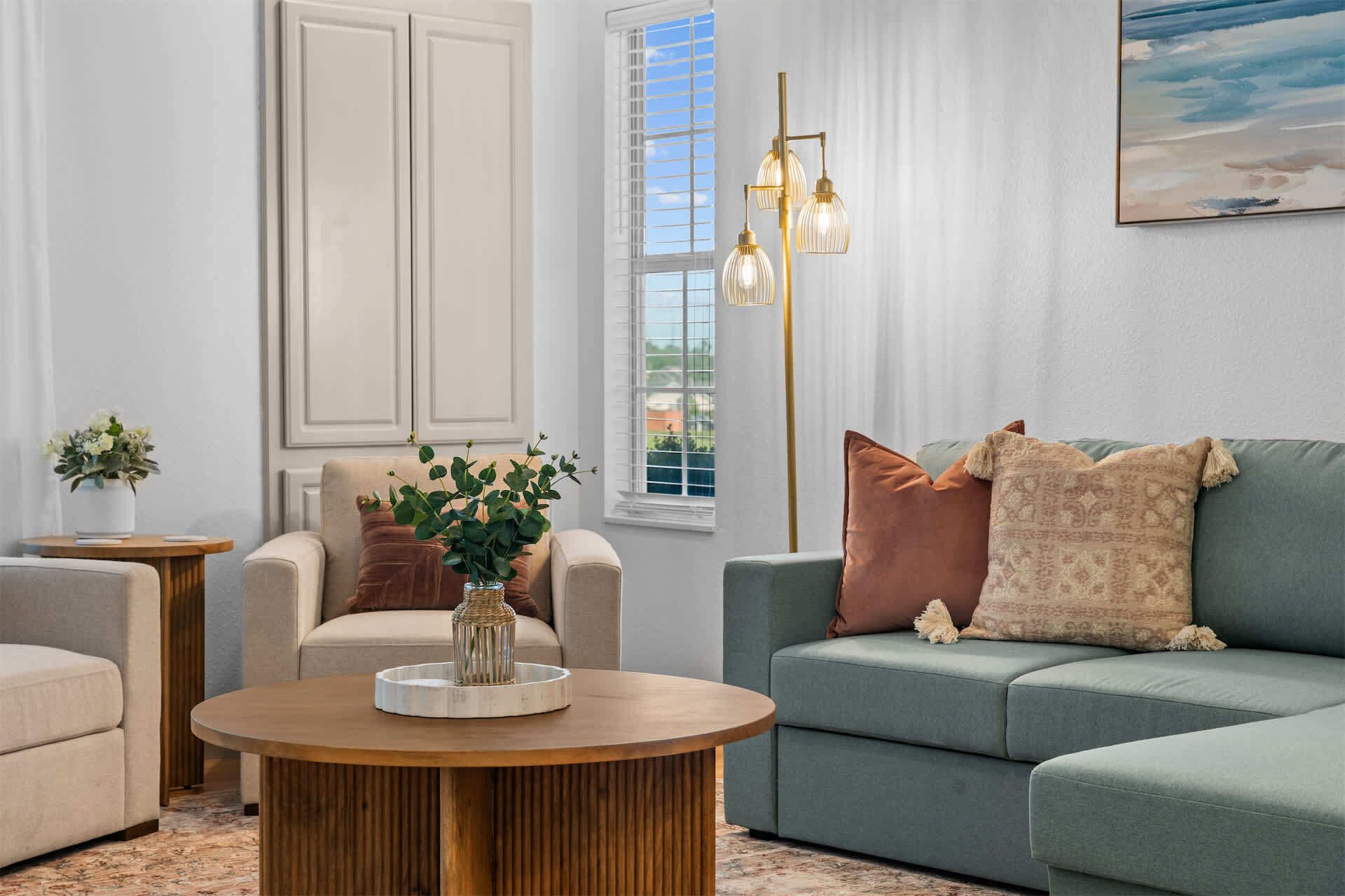 A living room with a sofa, armchairs, a coffee table, a vase with flowers, a painting, and a window with blinds.