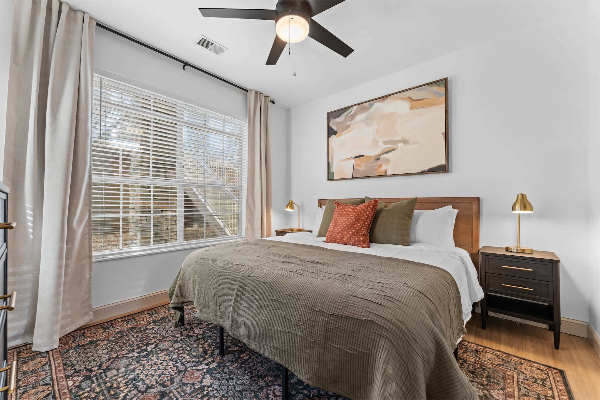 A bedroom with a bed, nightstands, a ceiling fan, a painting, and a window with blinds.