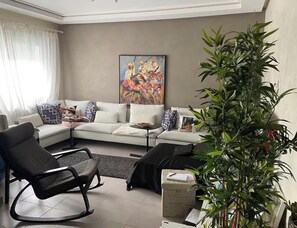 A living room with a sofa, a black rocking chair, a potted plant, and a painting on the wall. - Elegant Apartment in a Secured Residence with Pool Near the Beach (Bouznika)
