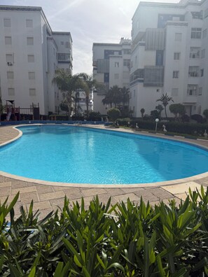 A swimming pool surrounded by buildings, with a playground and palm trees. - Elegant Apartment in a Secured Residence with Pool Near the Beach (Bouznika)