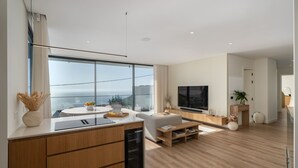 A modern kitchen with a large window, a dining area with a white table, and a living room with a TV and seating. - Superb modern villa with pool in Calheta|Amani (Arco Da Calheta)