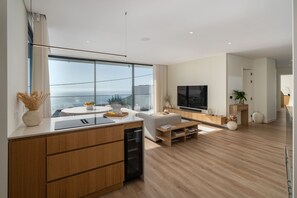 Eine moderne Küche mit großem Fenster, eine Essecke mit weißem Tisch und ein Wohnzimmer mit Fernseher und Sitzgelegenheiten.