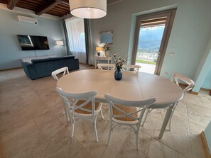 Zona pranzo con tavolo rotondo e sedie bianche, divano blu, televisione e vista panoramica attraverso un'ampia finestra.