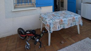 Een huis met een blauwe deur, een raam met een metalen traliewerk en een tafel met een tafelkleed met dessin.