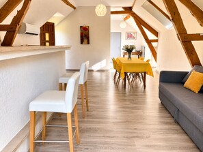Une chambre moderne de style loft, avec des poutres en bois apparentes, un coin repas agrémenté d’une nappe jaune et un canapé gris avec un coussin jaune.
