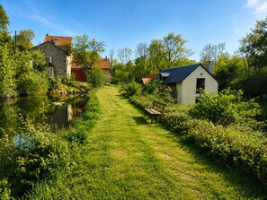 Un étroit sentier mène  à un petit bâtiment doté d’un toit en pente, entouré d’une végétation luxuriante et bordé d’une étendue d’eau paisible.