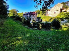 Un bâtiment rustique en pierre, doté d’un toit en pente, entouré d’une végétation luxuriante et surplombé d’un ciel bleu clair.