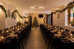 Une longue table à manger entourée de chaises, décorée de guirlandes lumineuses et de décorations de Noël, un sapin de Noël et une horloge au mur.