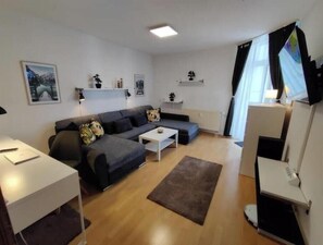 Ein modernes Wohnzimmer mit einem grauen Sofa, einem weißen Couchtisch und einem braunen Teppich. An den Wänden befinden sich Regale mit Dekoration und ein gerahmtes Bild.