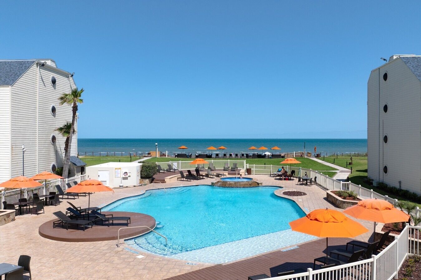 A pool area with lounge chairs and umbrellas, a view of the ocean, and a building with a white facade.