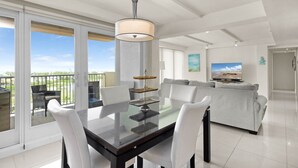 Een moderne woonkamer met een glazen eettafel, witte stoelen, een grijze bank, een televisie en een balkon met uitzicht.