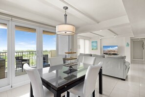 A modern living room with a glass dining table, white chairs, a grey sofa, a TV, and a balcony with a view. - Solare Seventh Floor Condo (South Padre Island)