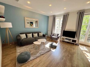 A living room with a sofa, two side tables, a television, and a large window with curtains. - Spacious, recently renovated apartment near the Louvre (Île-de-France)