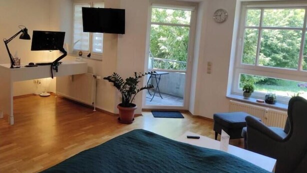 Huoneessa on sänky, työpöytä, jossa on tietokone, televisio, ikkuna, josta on näkymä vehreään maisemaan, ja parveke.