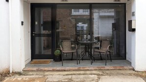 Une porte vitrée donne accès à un bâtiment, devant lequel se trouvent une petite table et deux chaises en osier.