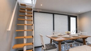 Un coin repas avec une table en bois dressée pour quatre personnes, un escalier avec des marches en bois et une fenêtre équipée de stores.