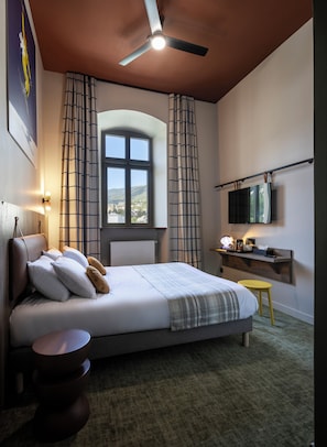Un dormitorio con cama, ventilador de techo, ventana con cortinas, mesita de noche y televisor montado.
