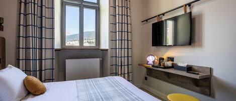 A hotel room with a bed, a desk with a flat-screen TV, a small stool, and a window with curtains.