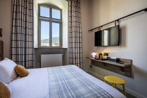 Habitación de hotel con cama, escritorio, televisor de pantalla plana, un taburete pequeño y ventana con cortinas.