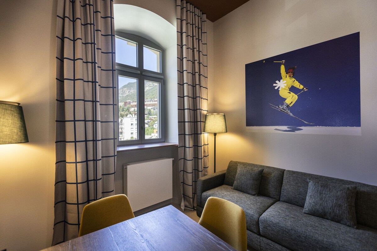 Habitació d'hotel amb taula de menjador, dues cadires, un sofà, una finestra amb cortines, una làmpada i un quadre penjat a la paret que representa un esquí.