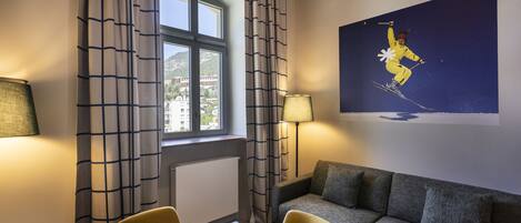 Une chambre d’hôtel comprenant une table à manger, deux chaises, un canapé, une fenêtre avec des rideaux, une lampe et un tableau représentant un skieur, accroché au mur.