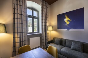 Habitación de hotel con una mesa de comedor, dos sillas, un sofá, una ventana con cortinas, una lámpara y un cuadro colgado en la pared de un esquiador.