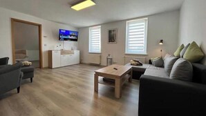 En moderne stue med sofa, sofabord, fjernsyn og et køkkenområde med skabe.