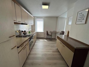 Eine kleine Küche mit Holzausstattung, ein Essbereich mit Tisch und Stühlen sowie ein Fenster mit Jalousien.
