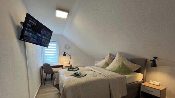 Et soveværelse med en seng, et skrivebord, en stol, et TV ophængt på væggen og et sengebord med en lampe.