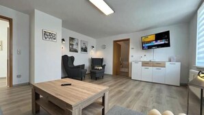 En moderne stue med et træbord, en grå sofa, et TV ophængt på væggen og et køkken med hvide skabe.