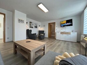 Ein modernes Wohnzimmer mit Couchtisch aus Holz, einer grauen Couch, einem an der Wand montierten Fernseher und einer Küchenzeile mit weißen Schränken.