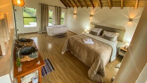 Een slaapkamer met een houten vloer, een bed met een beige sprei, een houten nachtkastje, een witte nachttafel met een lamp, en een raam met gordijnen.