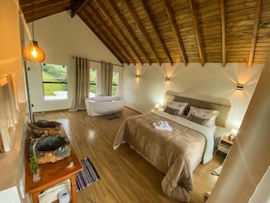 A bedroom with a wooden ceiling, a bed, a nightstand, a bedside table, a window, and a bathroom with a bathtub.