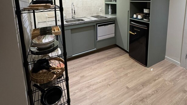 Een kleine keuken met een magnetron, oven en spoelbak. Er is een rek met servies en een pan aan de muur.