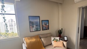 A modern living room with a sofa, a window with a view of buildings, and two framed pictures on the wall.