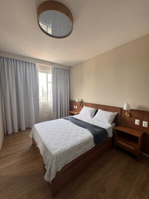 Ein Hotelzimmer mit einem Bett, einem Holz-Kopfteil, zwei Nachttischen, einem runden Spiegel und einem Fenster mit Vorhängen.