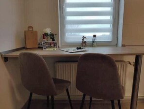 A room with a desk, two chairs, a radiator, and a window with blinds. - Feel-good apartment in the heart of Clausthal (Clausthal-Zellerfeld)