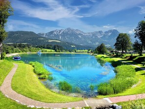 A clear lake with a pathway, surrounded by lush greenery and mountains in the background.