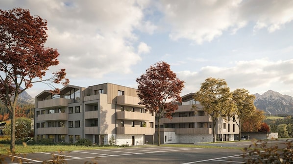 Un immeuble moderne à plusieurs étages, doté de balcons, entouré d’arbres et avec des montagnes en arrière-plan.