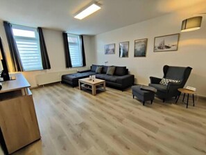 Ein modernes Wohnzimmer mit einer Couch, Sesseln, einem Couchtisch und einem kleinen Beistelltisch. Vier Fenster mit Vorhängen, eine Heizung und ein Wandbild sind zu sehen.