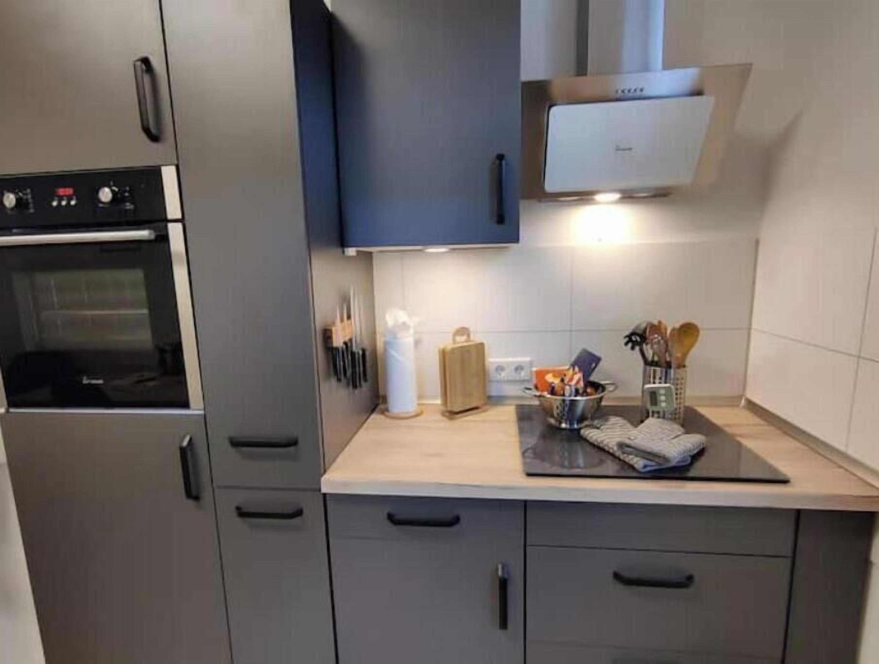 A modern kitchen with stainless steel appliances, a built-in oven, and a microwave. There are wooden cabinets and a countertop with a kettle and a basket of utensils.