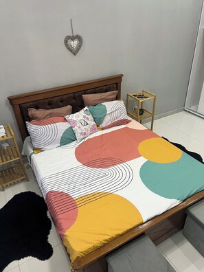 Een bed met een bedsprei met een patroon, een hartvormige decoratie aan de muur, en een houten nachtkastje met een lamp.